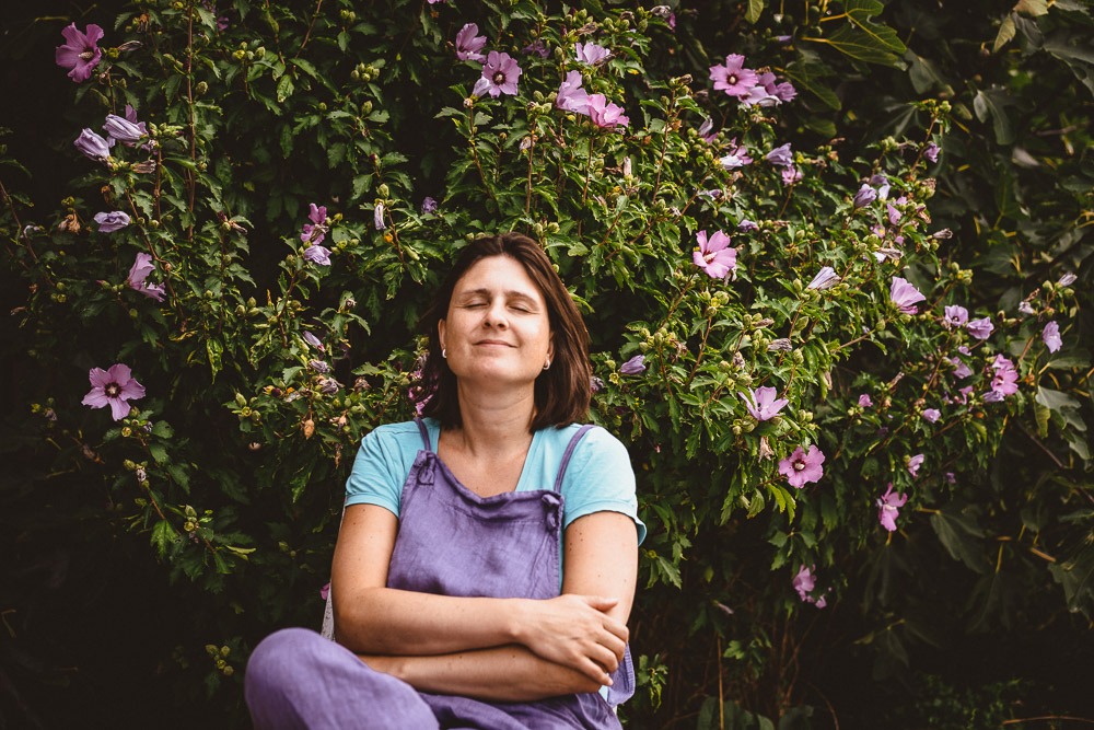 Selbstportrait Familienotografin mit lila Hose und sinnlichem Blick entspannt vor lila Blumenstrauch