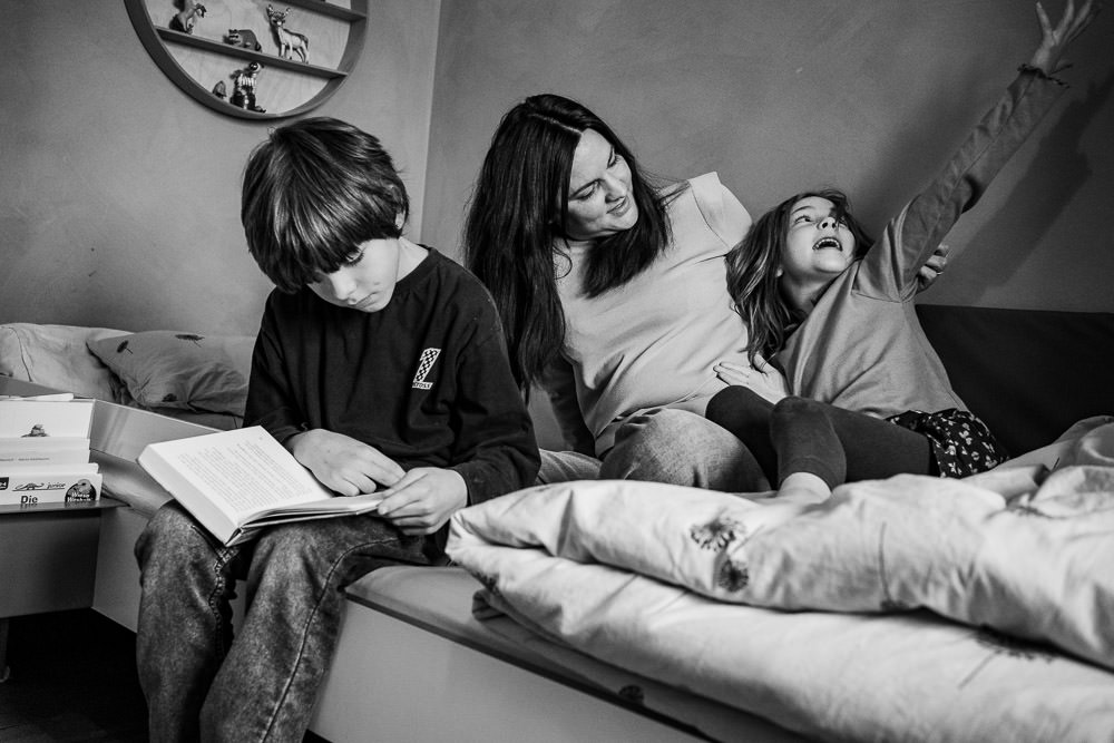 Familienfotos ungestellt bei dem die Mutter mit Kinder am Bett tobt und ein anderes Kind liest daneben